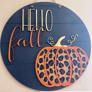 以下が含まれることがあります： 「Hello Fall」という金とオレンジの文字が書かれた丸い木製の看板。看板の右側には、ヒョウ柄のオレンジ色の大きなカボチャがあります。