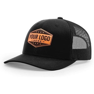 May include: Black trucker hat with a black mesh back. The front of the hat features a brown leather patch with the text "YOUR LOGO - HERE -" in white.
