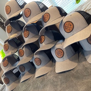 May include: A stack of gray and black trucker hats with a brown leather patch featuring the letters "I/S" and a cross design.