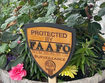 Può includere: Un cartello di legno con un bordo nero a forma di scudo. Il cartello recita "Protected by F.A.A.F.O. Fuck Around And Find Out Surveillance". Il cartello ha anche un teschio e ossa incrociate con una bandiera americana sullo sfondo.