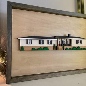 May include: A framed wood wall art piece featuring a 3D model of a white house with a brown roof and green landscaping. The house has multiple windows and a front porch.