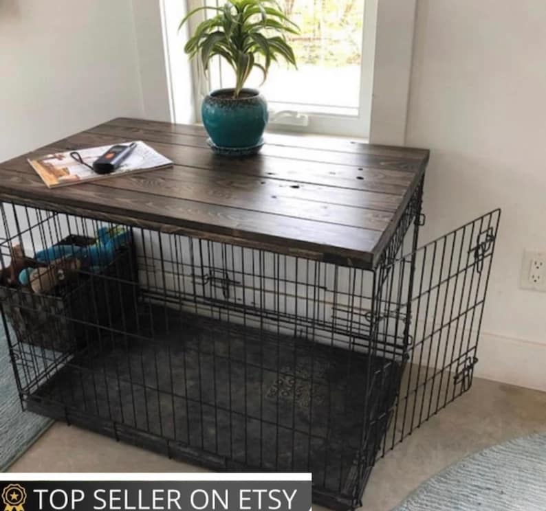 May include: A black wire dog crate with a dark brown stained wooden top. The crate features a hinged door on the right side and a black plastic tray at the bottom. A small green potted plant sits on the wooden surface next to a magazine.