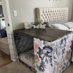 May include: A black wire dog crate with a dark wood top and a floral fabric cover featuring pink, grey, and white watercolor flowers. The crate has an open front door and contains a grey patterned cushion inside, with decorative items on top.