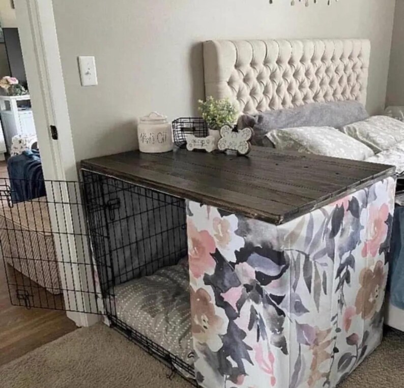 May include: A black wire dog crate with a dark wood top and a floral fabric cover featuring pink, grey, and white watercolor flowers. The crate has an open front door and contains a grey patterned cushion inside, with decorative items on top.