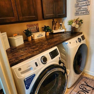 May include: A white laundry room with a wooden countertop and two white washing machines. The countertop has a sign that says "Laundry Room" and a sign that says "Lost Socks". There are also some plants on the countertop.