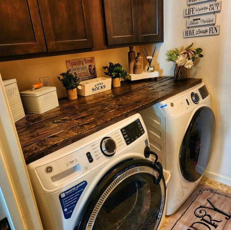 May include: A white laundry room with a wooden countertop and two white washing machines. The countertop has a sign that says "Laundry Room" and a sign that says "Lost Socks". There are also some plants on the countertop.