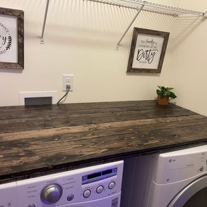 May include: A dark wood countertop with a white washer and dryer underneath. The countertop is in a laundry room with white walls and a white wire shelf above. There are two framed signs on the wall, one that says "The Laundry Room Do It Yourself" and the other that says "The laundry is looking at me Dirty Again".