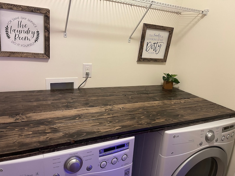 May include: A dark wood countertop with a white washer and dryer underneath. The countertop is in a laundry room with white walls and a white wire shelf above. There are two framed signs on the wall, one that says "The Laundry Room Do It Yourself" and the other that says "The laundry is looking at me Dirty Again".