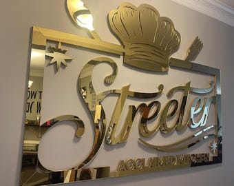 May include: Gold mirrored sign with the text "Streeter Acclaimed Kitchen" in a stylized font. A chef's hat and a spoon are depicted above the text.