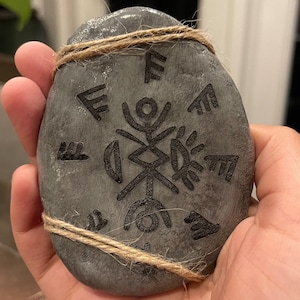 May include: A gray stone with a black design of a symbol with a circle in the center and lines radiating out. The stone is tied with twine.