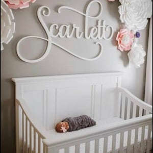 May include: A white wooden sign with the name "Scarlett" in cursive script hangs above a white crib with a baby sleeping inside. The crib is decorated with white paper flowers.