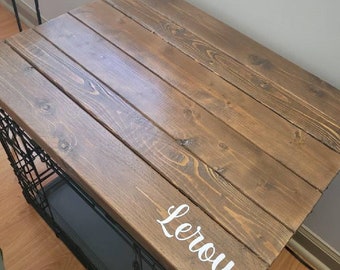 Può includere: Una cassa di legno con un top tinto di marrone e il nome "Leroy" scritto in lettere bianche. La cassa è appoggiata su una gabbia per cani in metallo nero.