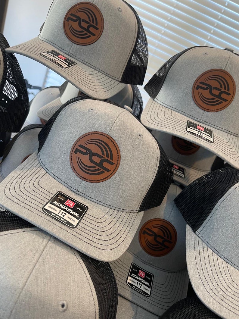 May include: A stack of gray Richardson 112 trucker hats with black mesh backs. Each hat features a brown leather patch with a circular logo. The hats are adjustable and have a structured front.