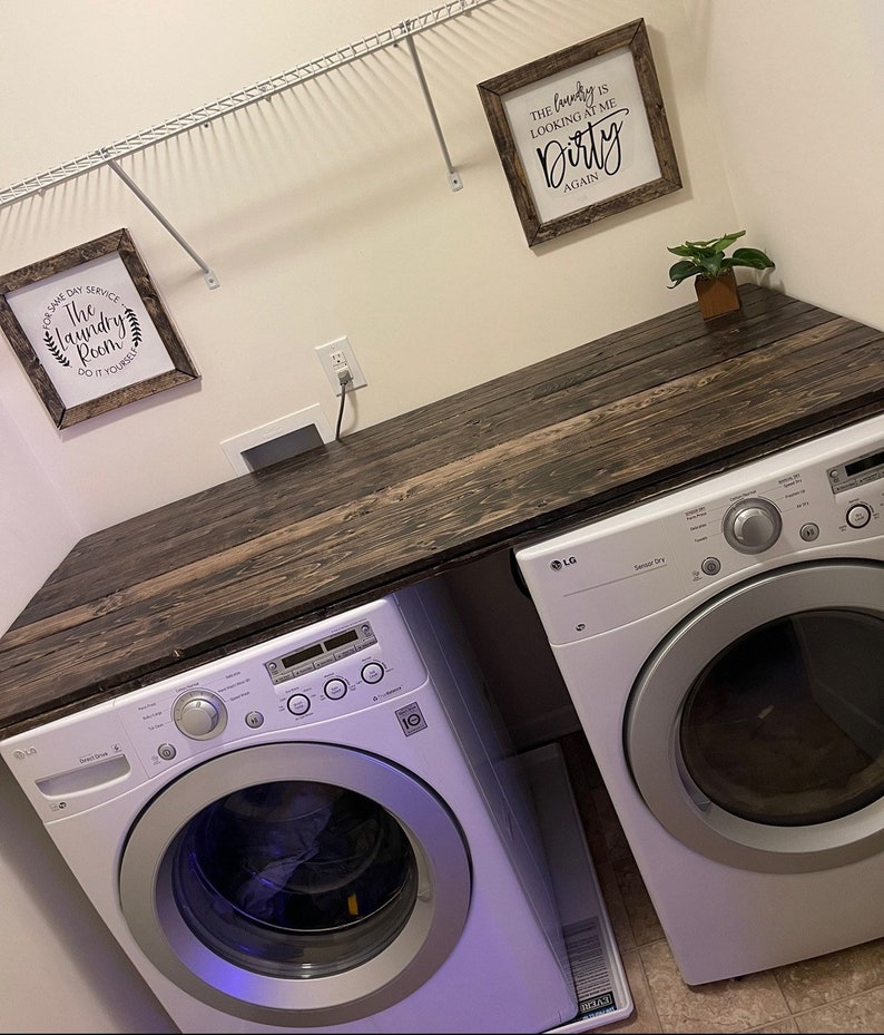 May include: A white laundry room with a wooden countertop and two white LG appliances, a washer and dryer. The countertop has two framed signs, one that says "The Laundry Room Do It Yourself" and the other that says "The Laundry Is Looking At Me Dirty Again".