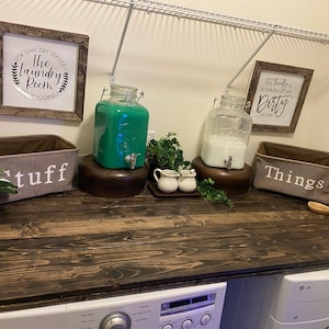 May include: A rustic wooden countertop with two large glass jars, one filled with green liquid and the other with white liquid. There are two smaller jars with white lids and a wooden spoon on the counter. Two fabric baskets with the words "Stuff" and "Things" are on the counter. The countertop is above a white washing machine and dryer.