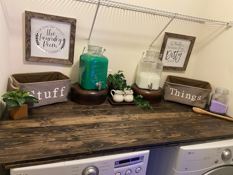 May include: A rustic wooden countertop with two large glass jars, one filled with green liquid and the other with white liquid. There are two smaller jars with white lids and a wooden spoon on the counter. Two fabric baskets with the words "Stuff" and "Things" are on the counter. The countertop is above a white washing machine and dryer.