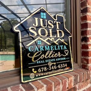 May include: A house-shaped black sign with gold and teal lettering. The text reads JUST SOLD, CARMELITA COLLIER, REAL ESTATE ADVISOR, 678-346-6334, and www.carmelitacollier.com. It features a gold key icon, a phone icon, and a globe icon.