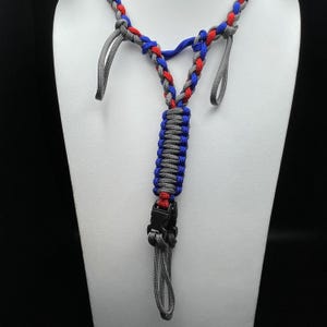 Pu&ograve; includere: Un cordino realizzato in paracord intrecciato nei colori rosso, blu e grigio. Il design presenta una sezione centrale piatta e spessa che si divide a forma di Y, con due anelli grigi in alto e una fibbia a sgancio rapido in plastica nera.