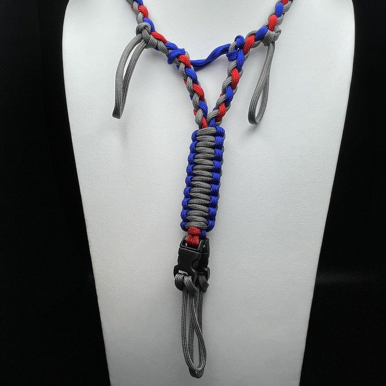 Pu&ograve; includere: Un cordino realizzato in paracord intrecciato nei colori rosso, blu e grigio. Il design presenta una sezione centrale piatta e spessa che si divide a forma di Y, con due anelli grigi in alto e una fibbia a sgancio rapido in plastica nera.