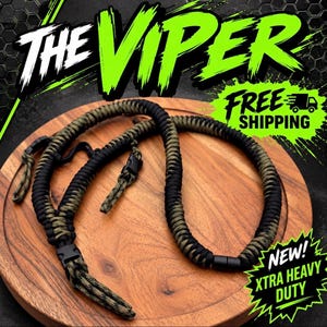 Può includere: Una tracolla per fotocamera in paracord intrecciato nero e verde oliva. La tracolla presenta fibbie laterali in plastica nera e connettori regolabili su entrambe le estremità. Il testo THE VIPER è visualizzato in alto, con NEW! XTRA HEAVY DUTY.