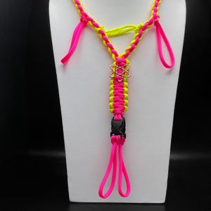 Pu&ograve; includere: Un'imbracatura in paracord rosa neon e giallo neon con un design intrecciato, un ciondolo centrale a forma di atomo color argento e una fibbia a sgancio laterale in plastica nera. L'accessorio include molteplici anelli e segmenti intrecciati.