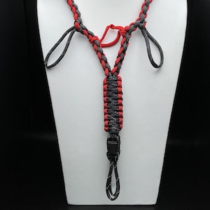 Pu&ograve; includere: Un cordino realizzato in paracord intrecciato rosso e nero con una sezione centrale tessuta, due anelli superiori e una fibbia in plastica nera nella parte inferiore. Il cordino presenta un motivo a punti riflettenti nei segmenti neri.