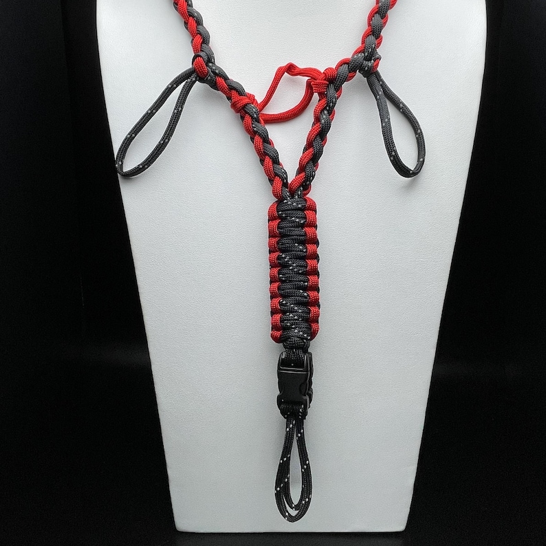 Pu&ograve; includere: Un cordino realizzato in paracord intrecciato rosso e nero con una sezione centrale tessuta, due anelli superiori e una fibbia in plastica nera nella parte inferiore. Il cordino presenta un motivo a punti riflettenti nei segmenti neri.