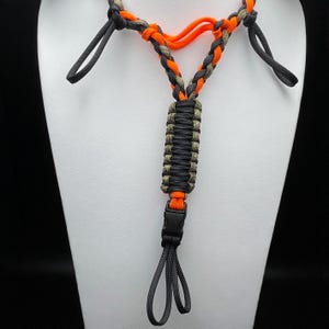 Pu&ograve; includere: Un cordino realizzato in paracord intrecciato nei colori arancione vivace, nero e verde oliva. Il design presenta una sezione centrale intrecciata, due anelli laterali e una fibbia in plastica nera sul fondo, che termina con un anello nero.