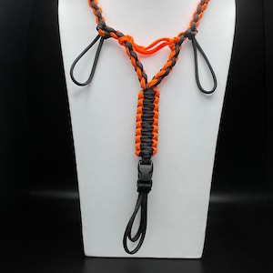 Pu&ograve; includere: Un cordino realizzato in paracord arancione e grigio scuro con un design intrecciato. L'accessorio include due anelli laterali, una sezione centrale intrecciata verticalmente e una fibbia a sgancio rapido in plastica nera con un anello.