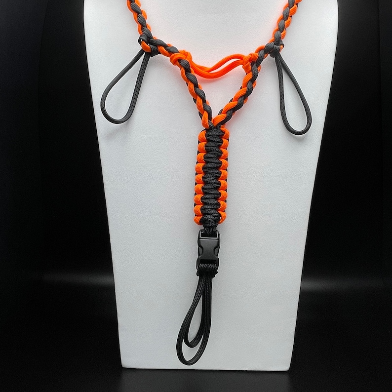 Pu&ograve; includere: Un cordino realizzato in paracord arancione e grigio scuro con un design intrecciato. L'accessorio include due anelli laterali, una sezione centrale intrecciata verticalmente e una fibbia a sgancio rapido in plastica nera con un anello.