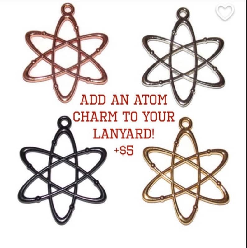 Pu&ograve; includere: Quattro ciondoli in metallo a forma di atomo con finiture in oro rosa, argento, nero e oro. Ogni ciondolo presenta un occhiello per l'aggancio. Il testo al centro recita: ADD AN ATOM CHARM TO YOUR LANYARD! +$5. Design di tipo scientifico.