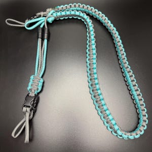 May include: A handmade lanyard in a turquoise and gray color scheme. The lanyard is made of braided cord and features a black plastic buckle and two black plastic connectors. The lanyard is designed to be worn around the neck.