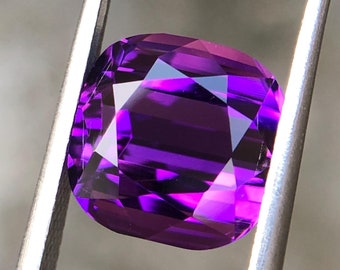 Könnte beinhalten: Ein tiefvioletter Edelstein mit einem Kissen-Schliff, gehalten von einer Pinzette. Der Edelstein hat eine hohe Klarheit und Brillanz.