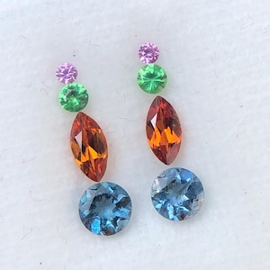 May include: A collection of colorful gemstones in a clear plastic case. The gemstones include round blue, oval orange, round green, and round pink stones. The gemstones are arranged in two vertical columns, with each column containing a variety of shapes and colors.