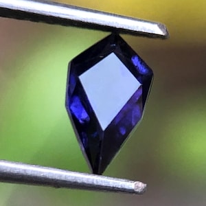 May include: A faceted, deep blue gemstone held by tweezers. The gem has a kite-like shape with a square facet at the top. The background is a soft blur of green and yellow.