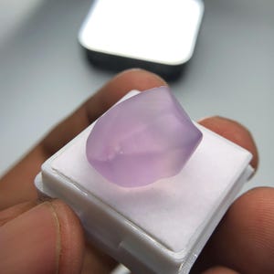 May include: A pale purple gemstone with a polished surface, set on a white plastic base. The gemstone is held in a person's hand. The background is blurred. The gemstone is likely a cabochon.