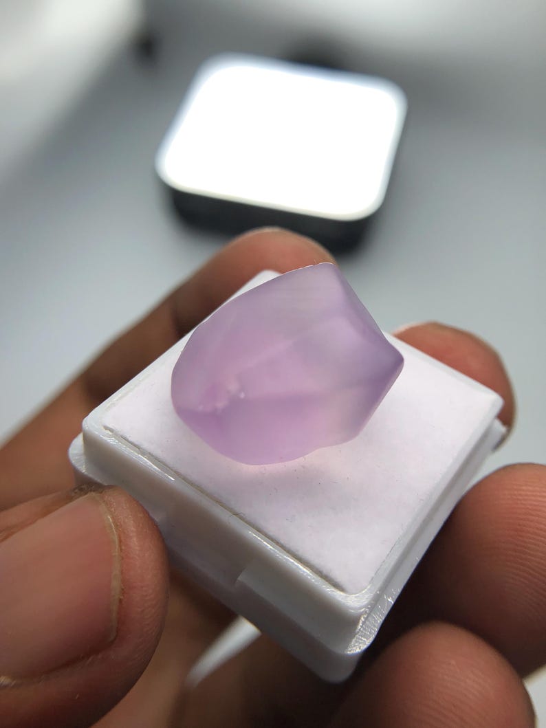 May include: A pale purple gemstone with a polished surface, set on a white plastic base. The gemstone is held in a person's hand. The background is blurred. The gemstone is likely a cabochon.
