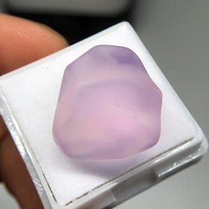 May include: A pale purple gemstone with a smooth, rounded form, presented in a white plastic display case. The stone has a soft, diffused appearance, with subtle colour variations. The gemstone is a natural, unpolished specimen.