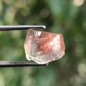 Puede incluir: Una gema facetada y translúcida con un interior marrón rojizo y un exterior claro y de color claro. La gema está sujeta por unas pinzas sobre un fondo verde borroso, mostrando su estructura cristalina natural. La gema parece ser una piedra solar.