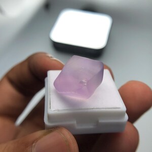May include: A pale purple gemstone, possibly amethyst, sits on a white display stand. The gemstone is rectangular with rounded edges. The background features a silver square object and a blurred white surface.