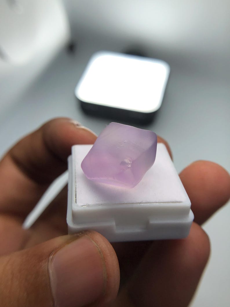 May include: A pale purple gemstone, possibly amethyst, sits on a white display stand. The gemstone is rectangular with rounded edges. The background features a silver square object and a blurred white surface.