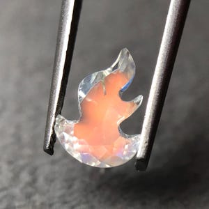 Könnte beinhalten: Ein klarer, facettierter Edelstein in Flammenform mit rosafarbenem Inneren. Der Edelstein wird von einer Pinzette gehalten.