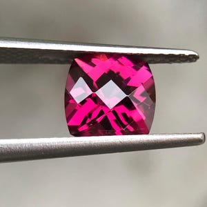 Könnte beinhalten: Ein lebendiger, kissenförmig geschliffener Edelstein in einem tiefen Magentaton. Die facettierte Oberfläche reflektiert das Licht und erzeugt einen funkelnden Effekt. Gehalten von einer silbernen Pinzette, ist der Edelstein quadratisch mit abgerundeten Ecken.
