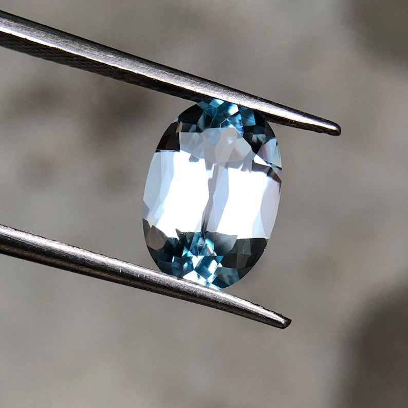 Op de afbeelding: Een gefacetteerde, ovaalvormige aquamarijn edelsteen, vastgehouden door een pincet. De edelsteen heeft een lichtblauwe kleur met interne reflecties. De pincet is gemaakt van zilverkleurig metaal. De achtergrond is een vage, neutrale tint.