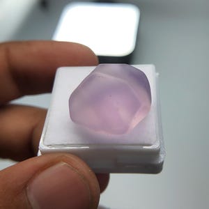 May include: A pale purple gemstone with a smooth, rounded form. The stone is presented on a small, white plastic tray. The image is well-lit, emphasising the stone's colour and texture.