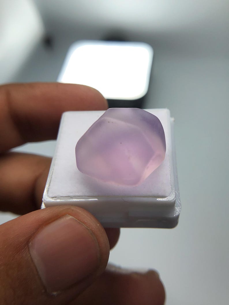 May include: A pale purple gemstone with a smooth, rounded form. The stone is presented on a small, white plastic tray. The image is well-lit, emphasising the stone's colour and texture.