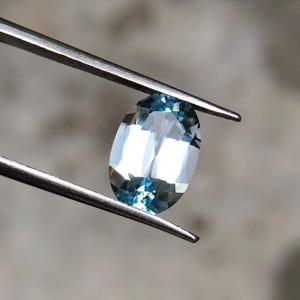 Op de afbeelding: Een ovaalvormige, lichtblauwe edelsteen, vastgehouden door een zilveren pincet. De gefacetteerde oppervlakte van de edelsteen reflecteert licht, wat een sprankelend effect cre&euml;ert. De kleur van de edelsteen is een bleek, doorschijnend blauw, met een vleugje aqua. De achtergrond is vaag en neutraal.