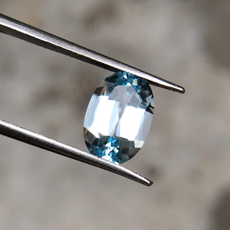 Op de afbeelding: Een ovaalvormige, lichtblauwe edelsteen, vastgehouden door een zilveren pincet. De gefacetteerde oppervlakte van de edelsteen reflecteert licht, wat een sprankelend effect cre&euml;ert. De kleur van de edelsteen is een bleek, doorschijnend blauw, met een vleugje aqua. De achtergrond is vaag en neutraal.