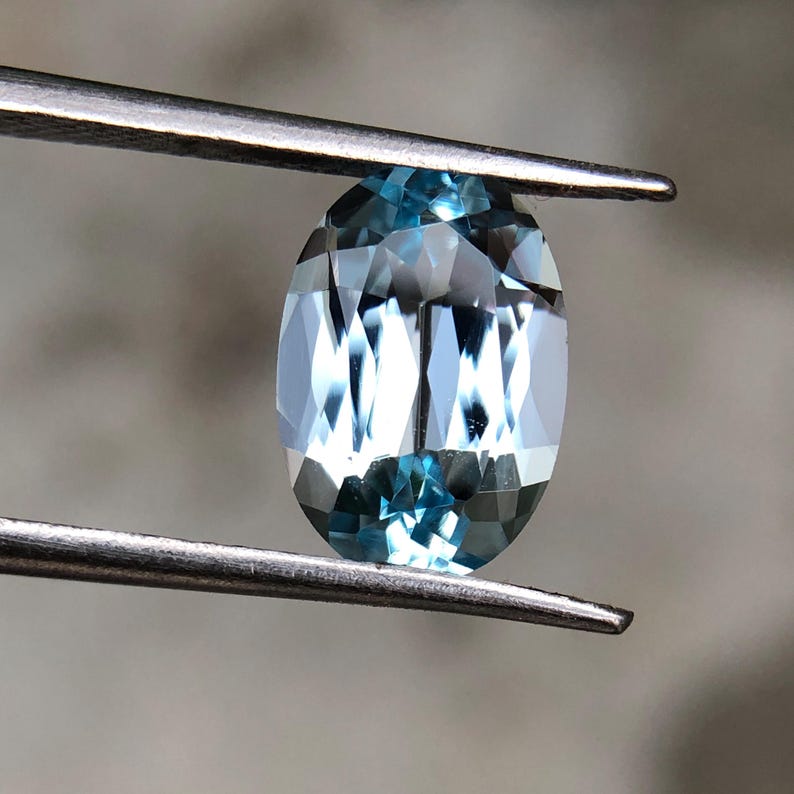Op de afbeelding: Een ovaalvormige, lichtblauwe edelsteen, vastgehouden door een pincet. De edelsteen heeft meerdere facetten die het licht reflecteren, waardoor een sprankelend effect ontstaat. De kleur van de steen varieert van lichtblauw tot dieper blauw, met een gepolijst oppervlak.