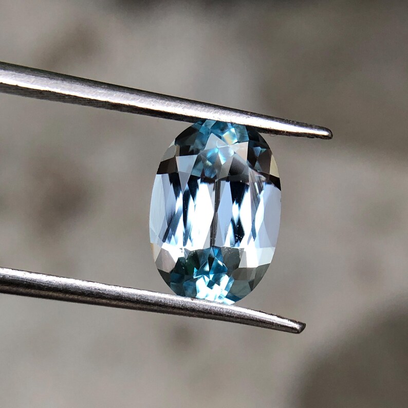 Op de afbeelding: Een ovaal geslepen, lichtblauwe edelsteen, vastgehouden door een metalen pincet. De edelsteen heeft meerdere facetten die het licht reflecteren, waardoor een sprankelend effect ontstaat. De edelsteen is lichtblauw van kleur, met een gepolijst oppervlak.